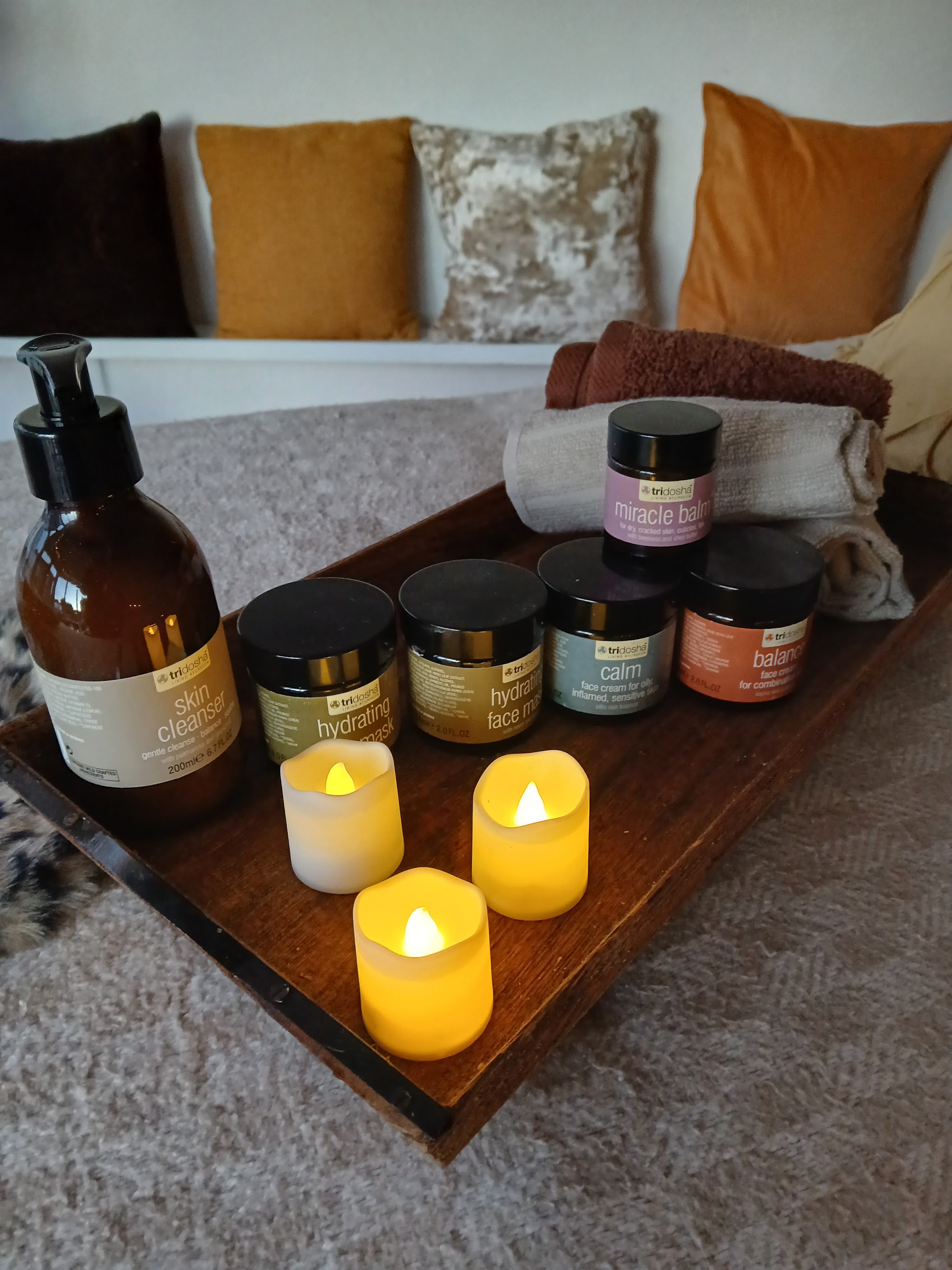 A tray of massage oils and creams with candles on the couch, mustard and gold cushions behind